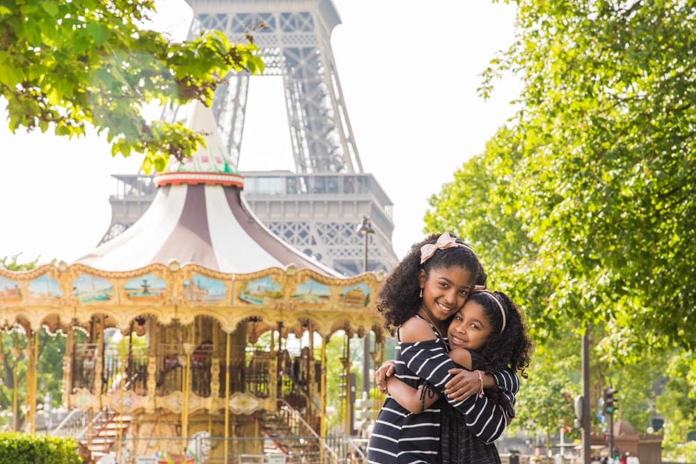 Two sisters of color embrace with Paris' iconic carousel and Eiffel tower behind them, one of the best places to travel with kids in Europe.