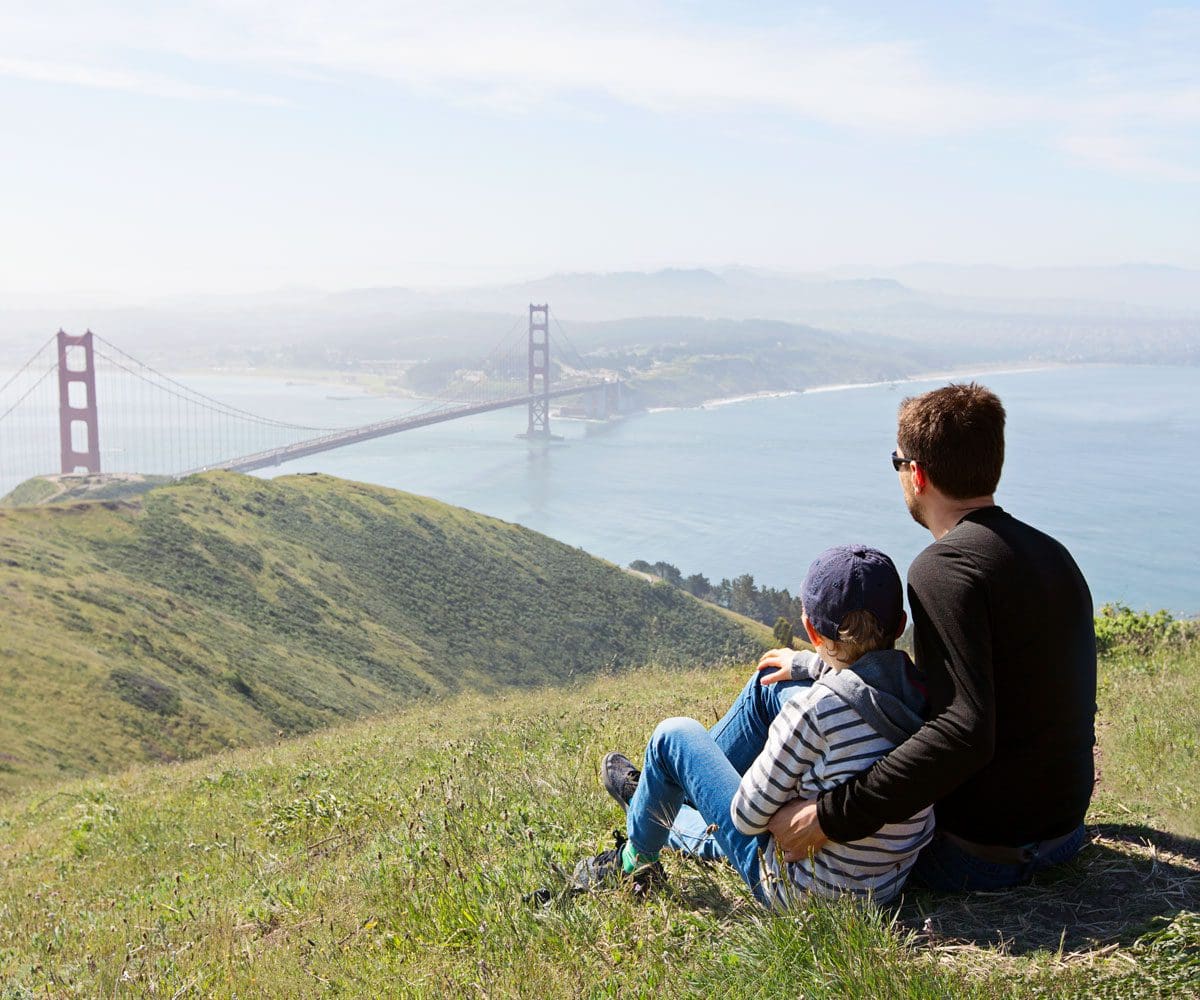 A dad and child sit together while looking over green hills toward the Golden Gate Bridge.
