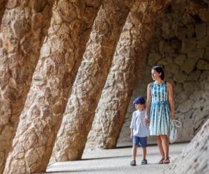 A mom holds the hand of her young son while they walk through a stone hallway in Barcelona.