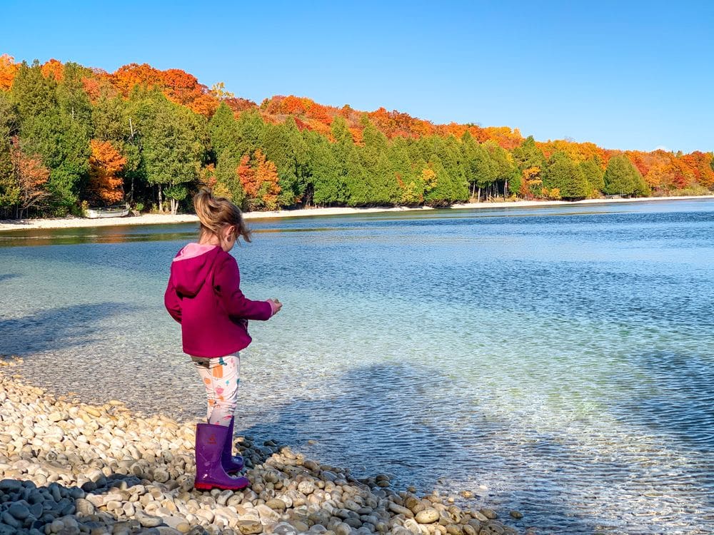 A young girl wearing a pink coat and purple rain boots stands at the edge of the water on School House Beach with an array of colorful fall foliage in the distance.