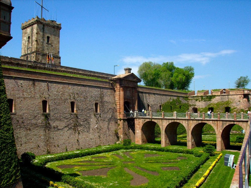 In the distance, several people meander across the stone bridge at Majestic Montjuic Castle, which is surrounded by green lawns and the stone castled behind them.