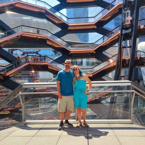 A man and woman stand together with the Vessel in Hudson Yards behind them.