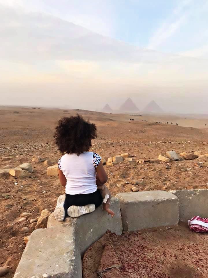A young African-American girl sits along a stone wall looking toward the three pyramids of Giza.