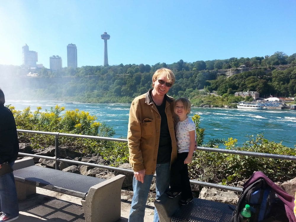 A dad and his daughter smile together above Niagara Falls with the Ontario skyline in the distance.