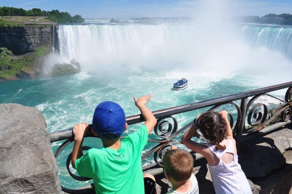 Three kids stand at a railing looking over it into Niagara Falls, where a boat is on the water headed toward the falls.