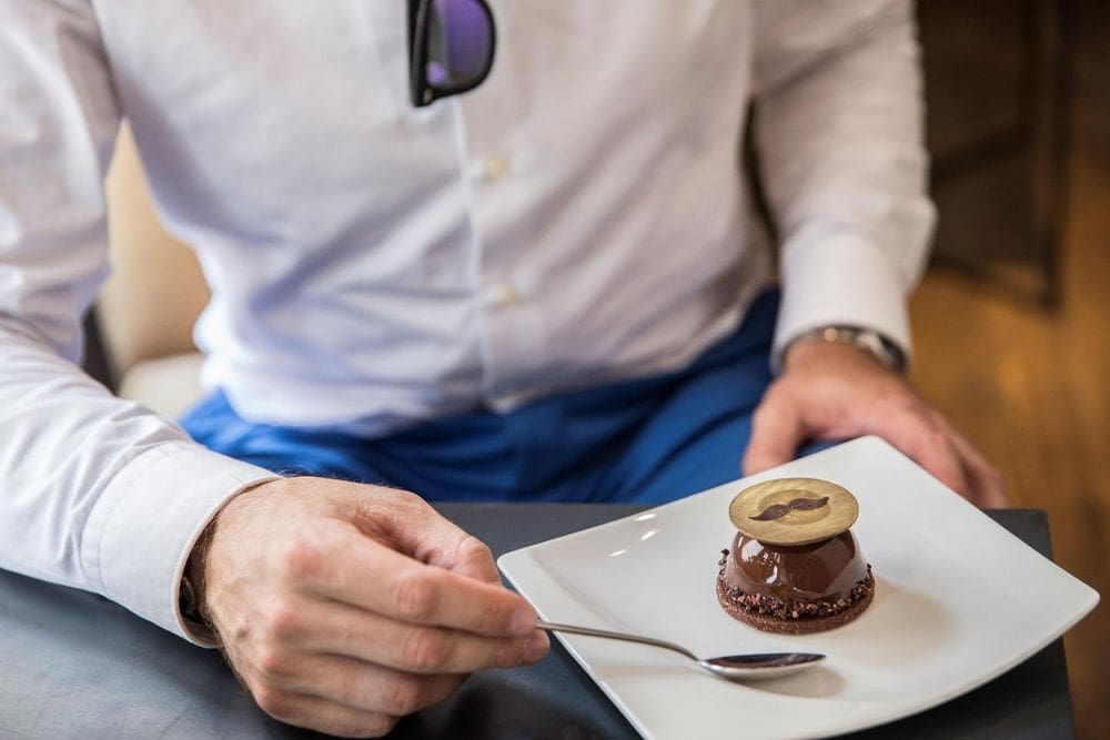 A close up of a gentleman's chest and arm reaching out for a delicate chocolate desert on a white plate at Un dimanche à Paris, one of the best dessert destinations in Paris for families.