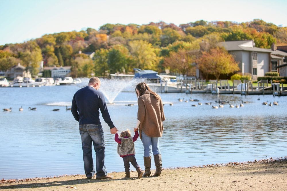 Two parents hold the hands of their young daughter on the shore of Lake Geneva, with brilliant fall colors in the distance.
