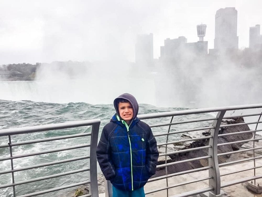A young boy wearing a blue jacket smiles with the mist of Niagara Falls behind him and a view of the Ontario skyline behind the mist.