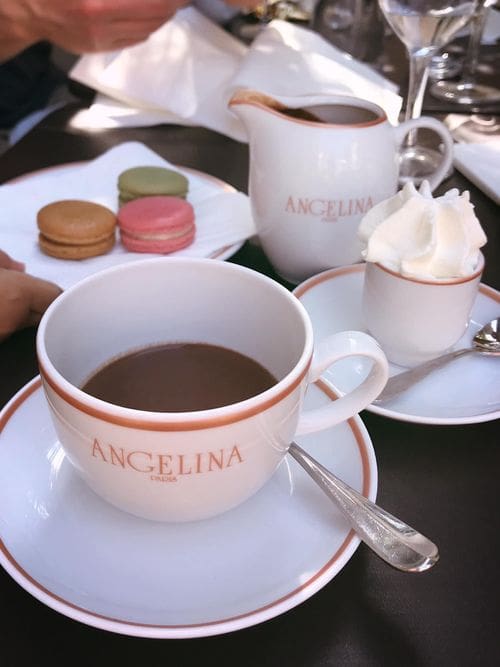 A Parisian table holds several macrons, a tea cup reading "Angelina", and other items for tea-time.