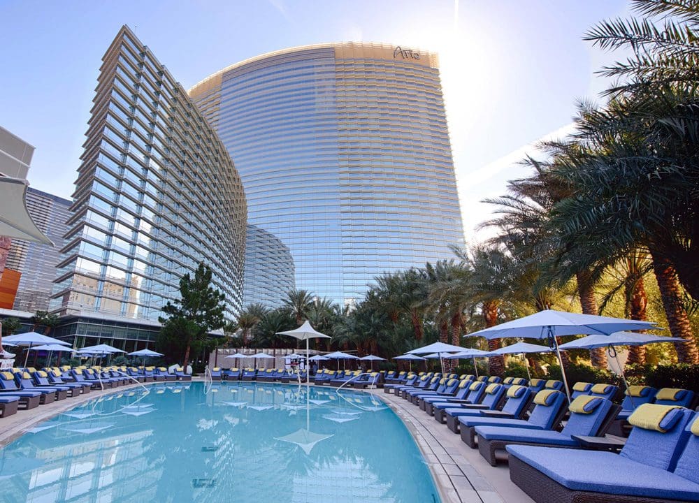 The pool and pool deck, featuring blue poolside loungers, at the ARIA Resort and Casino. The resort buildings are in the distance.