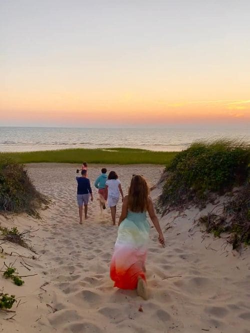 Several people walk along a sandy beach between dunes toward the water at sunset on Cape Cod.
