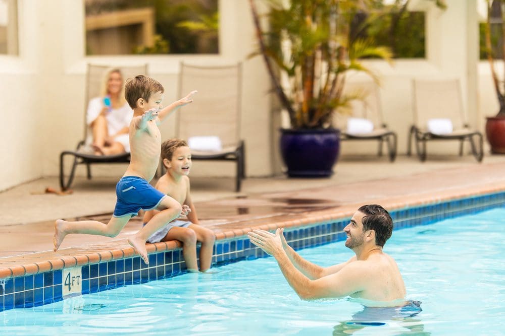 A young boy jumps into his dads arms, who is standing in the pool at the Bahia Resort Hotel, while mom and brother look on. This is one of the best home bases for your San Diego itinerary with kids.