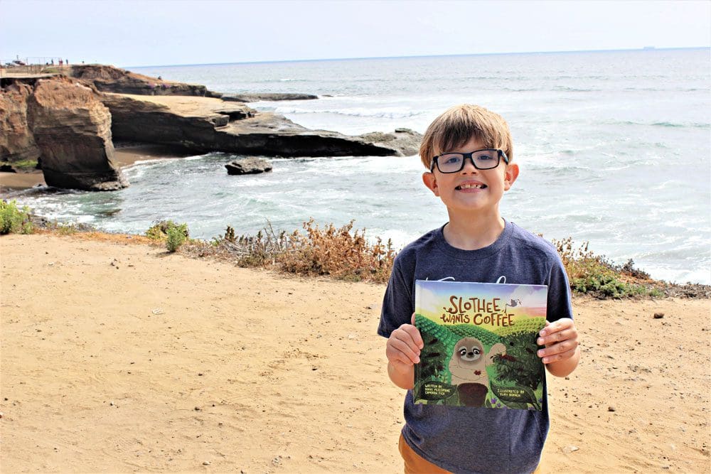 A young boy stands on the shore near San Diego holding a book titled "Slothie Wants Coffee".