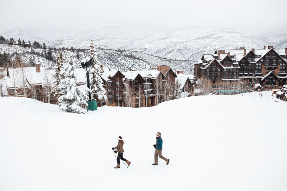 Two people walk across the snow, with the expansive resort and grounds of the The Ritz-Carlton, Bachelor Gulch behind them.