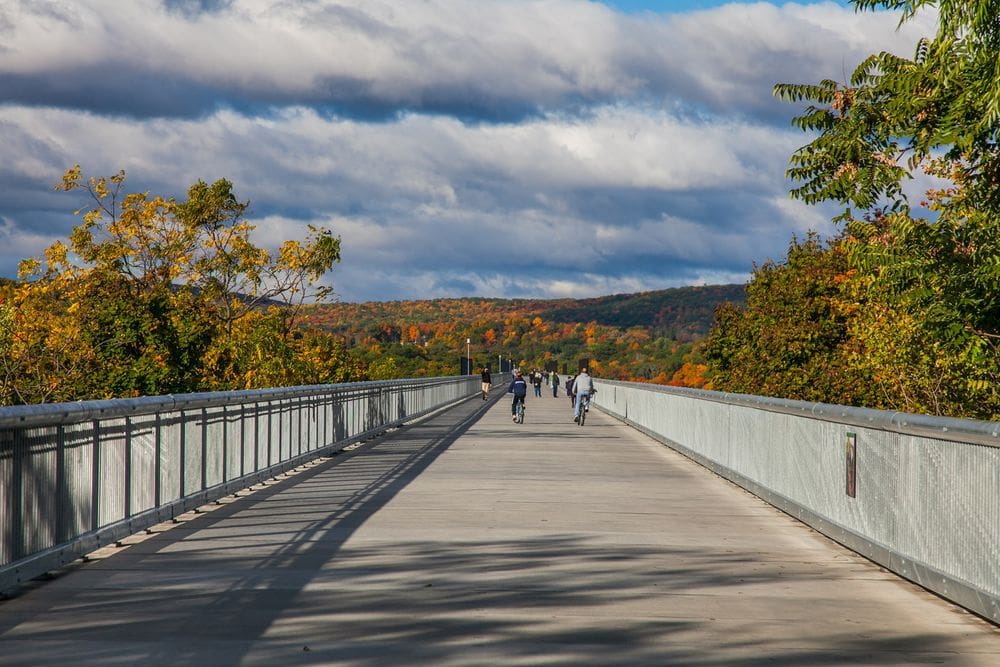 Several bikers and pedestrians cross the Walkway Over the Hudson State Historic Park amidst brilliant fall foliage.
