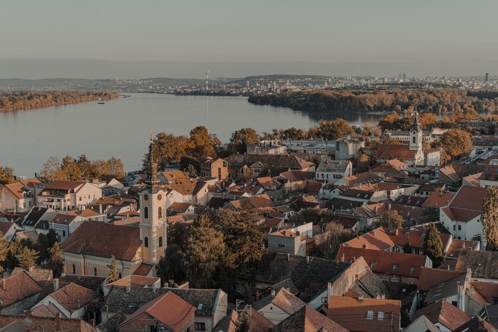 An aerial view of Belgrade, Serbia, featuring it's charming town, church steeple, and pond.