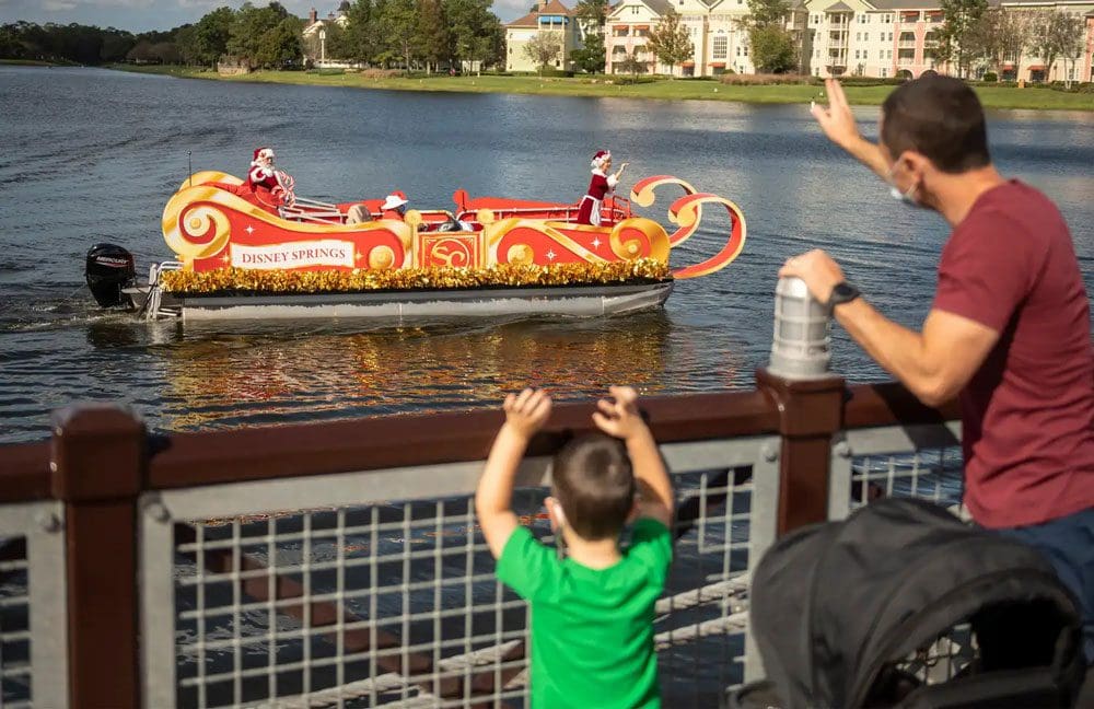 A father and son standing on a bridge wave to Santa and Mrs. Clause as they pass by on a holiday float near Disney Springs, while celebrating the holidays in Disney as a family.