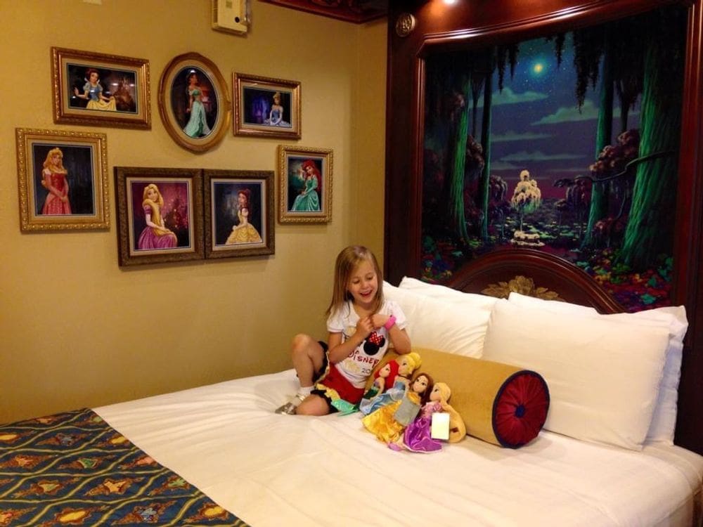 A young girl sits on a bed at Disney's Port Orleans - Riverside Resort, one of the best Disney moderate resorts for families, surrounded by plush Disney princess dolls.