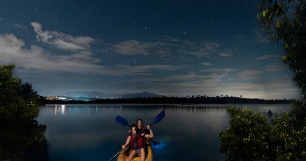 Two people paddle along in a canoe looking for bioluminescent life in Laguna Grande, with a view of city lights in the distance, one of the best things to do in Puerto Rico with kids.