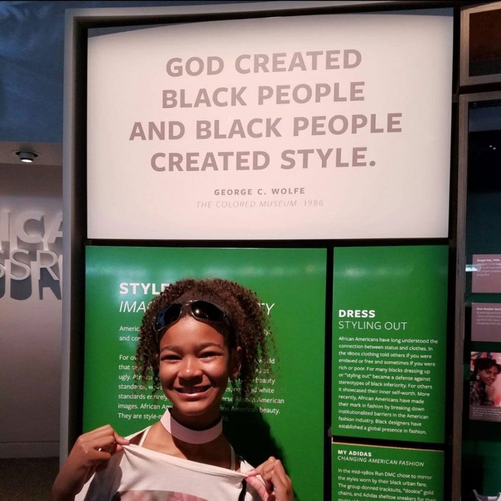A young African-American teen shows off her shirt in front of a sign that reads "God Created Black People and Black People Created Style" at the National Museum of African American History and Culture.