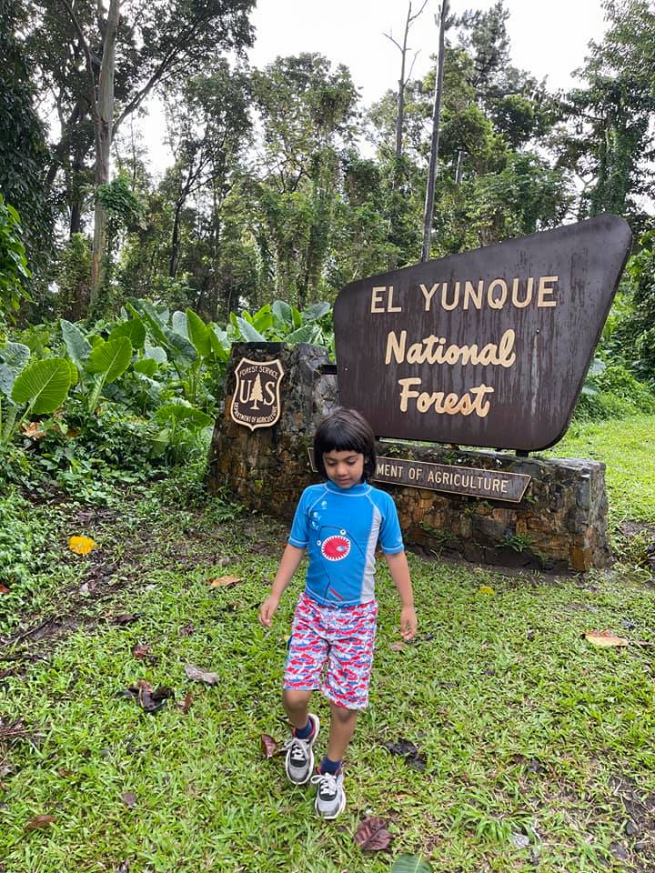 A young boy stands in front of a sign for El Yunque National Forrest, with lush greenery around him.