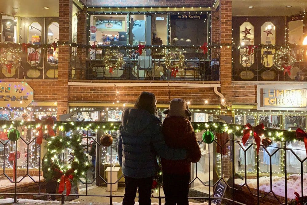 Two kids in winter gear stand together looking out onto the holiday decorations in Breckenridge. 