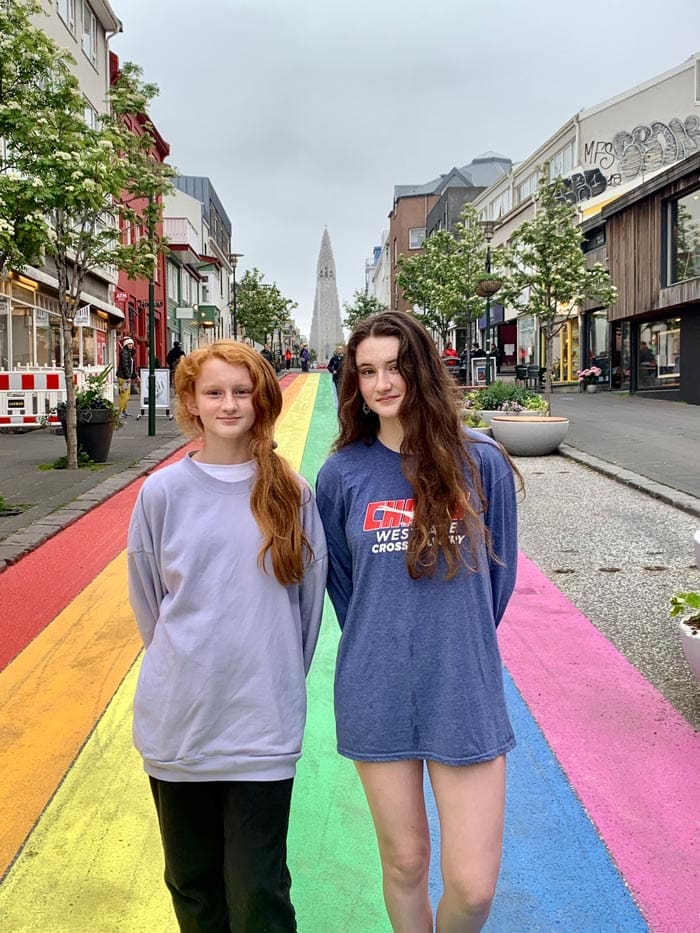 Two girls stand together with a view of Reykjavik in the distance. The street is painted like a rainbow.
