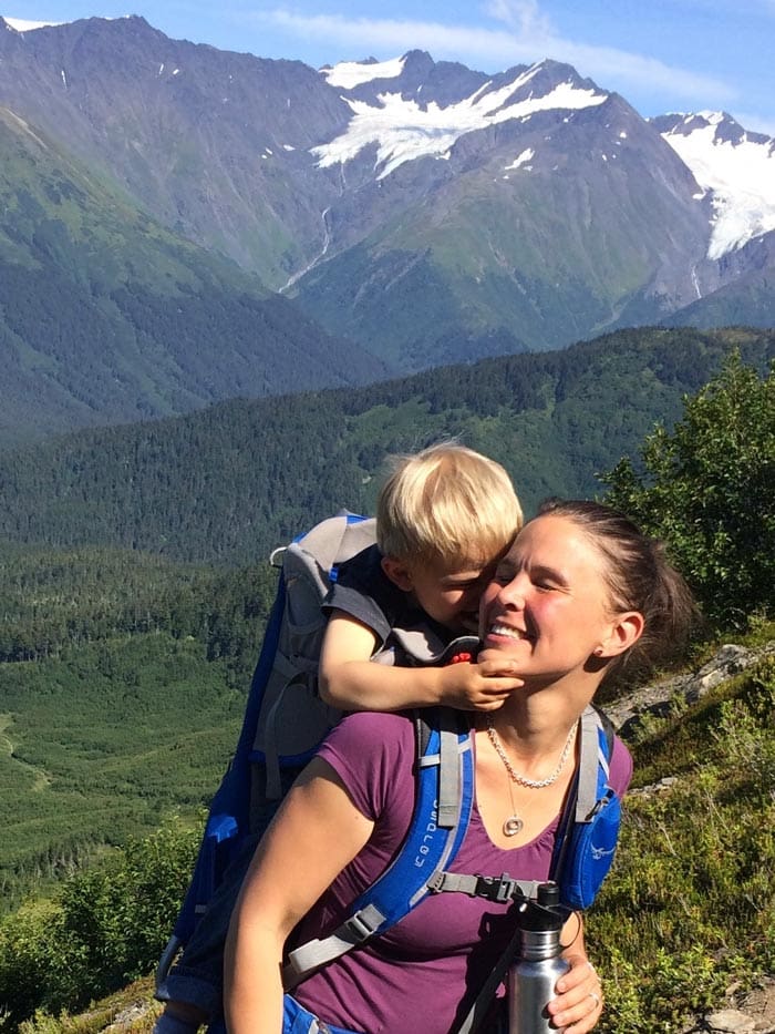 A mom carriers her baby on her back using a backpack-style carrier, the baby reached up toward the mother to give her a snuggle, while hiking in the mountains.