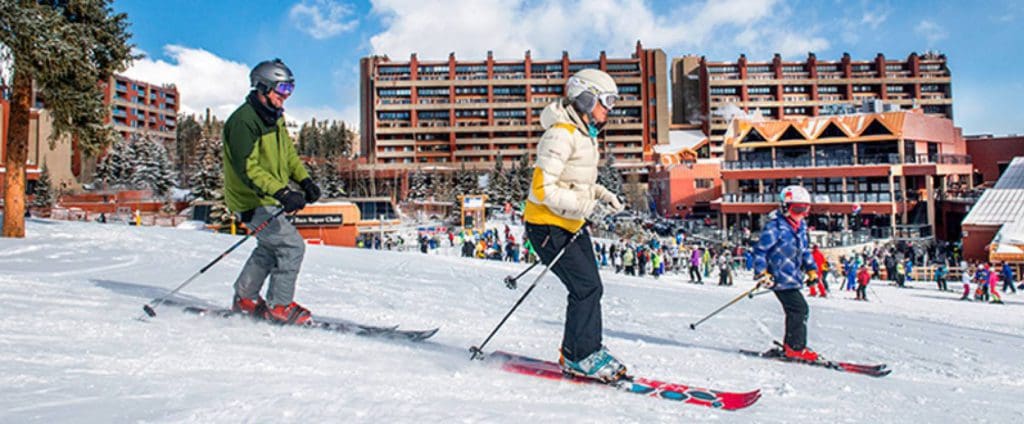A family of three skis down a slope with a view of Beaver Run Resort & Conference Center behind them, one of the best places to stay in Breckenridge with kids this winter.