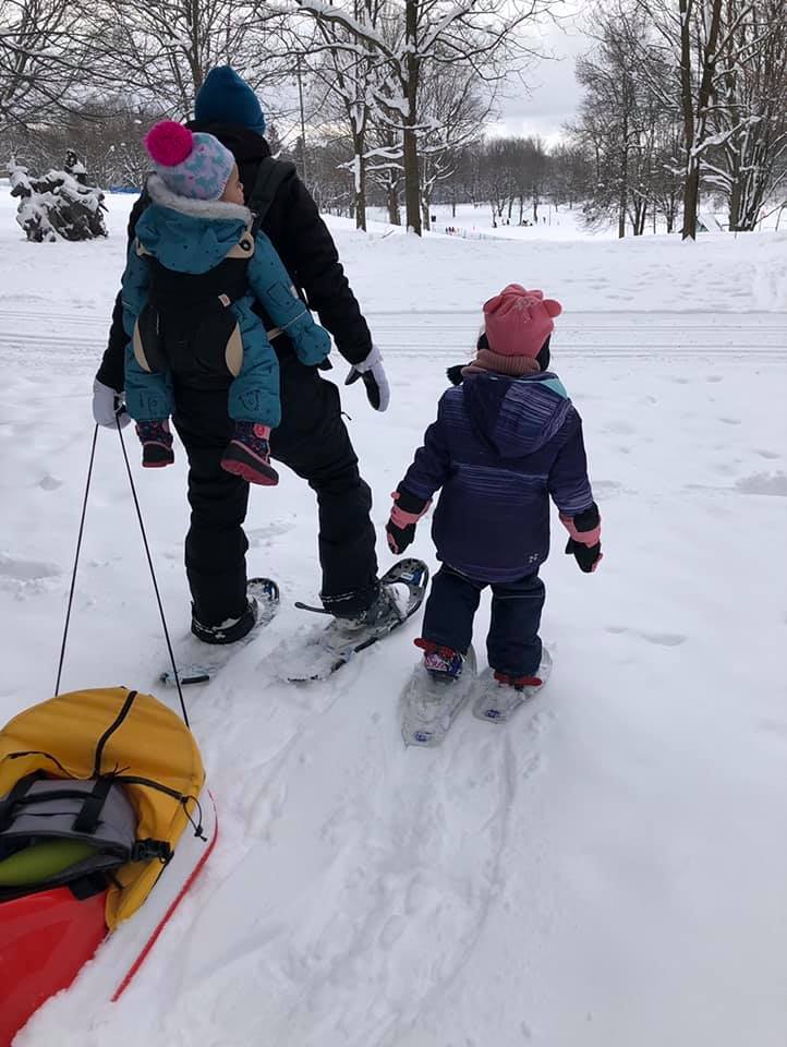 A mom carrier her child on her back using a baby carrier, while pulling a sled and snowshoeing with her other child.