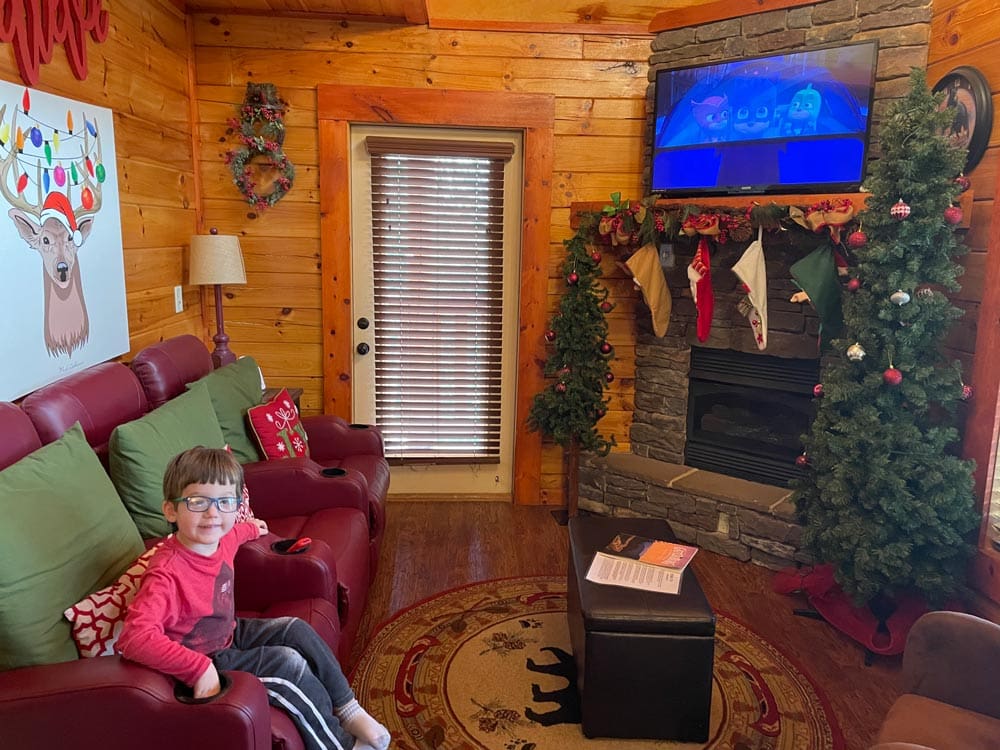A young boy sits on a couch inside a cozy cabin at Elk Springs during the Christmas holiday, a tree and stockings can be seen across the wood-paneled room.