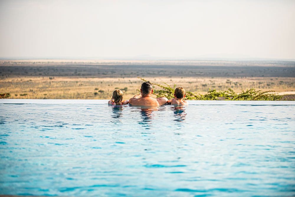 A dad and his two kids look out onto the Western Serengeti from a hotel pool.