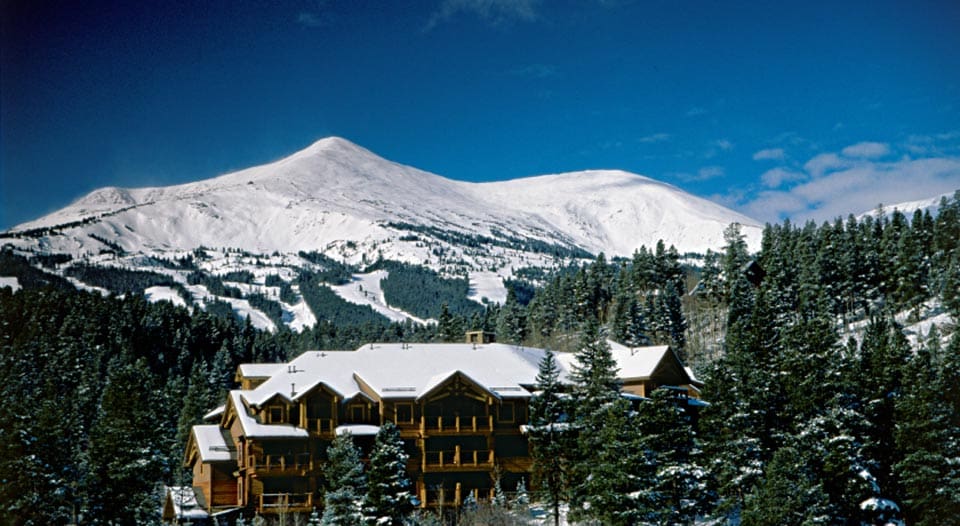 A view of one of the resort buildings at the Mountain Thunder Lodge through the pines. Behind the building is snow-covered mountain.
