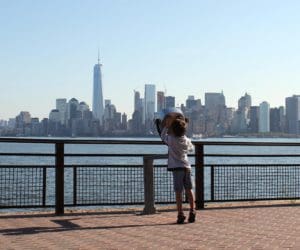 A young child leans up to look through a observation lens on the edge of a river, across from which is the NYC skyline.