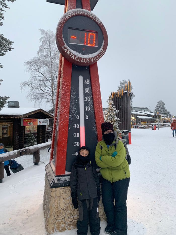 A mom and her son stand under a sign reading "-10" while visiting Rovaniemi in the winter.