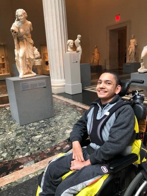 A teen boy, who uses a wheel chair, is sitting next to a sculpture at the Met.