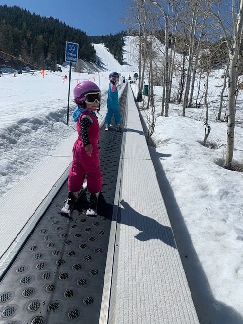 Two young kids in full snow gear ride the magic carpet up a ski slope.