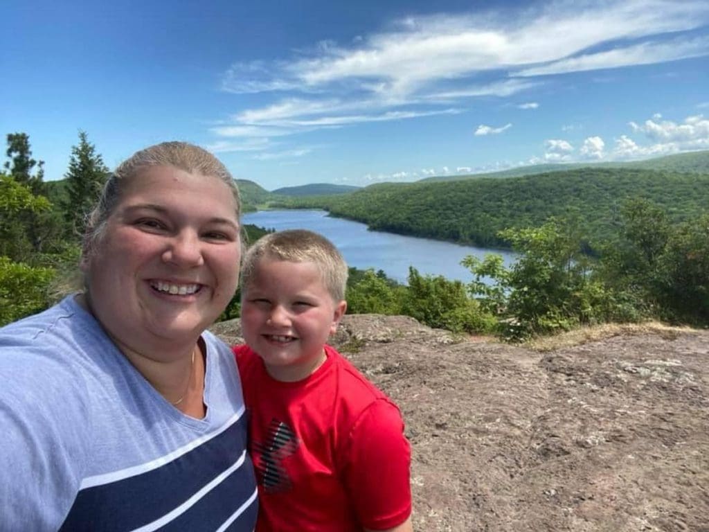 A mom and her son smile after hiking to the top of an overlook, with a view of the Lake of the Clouds behind them.