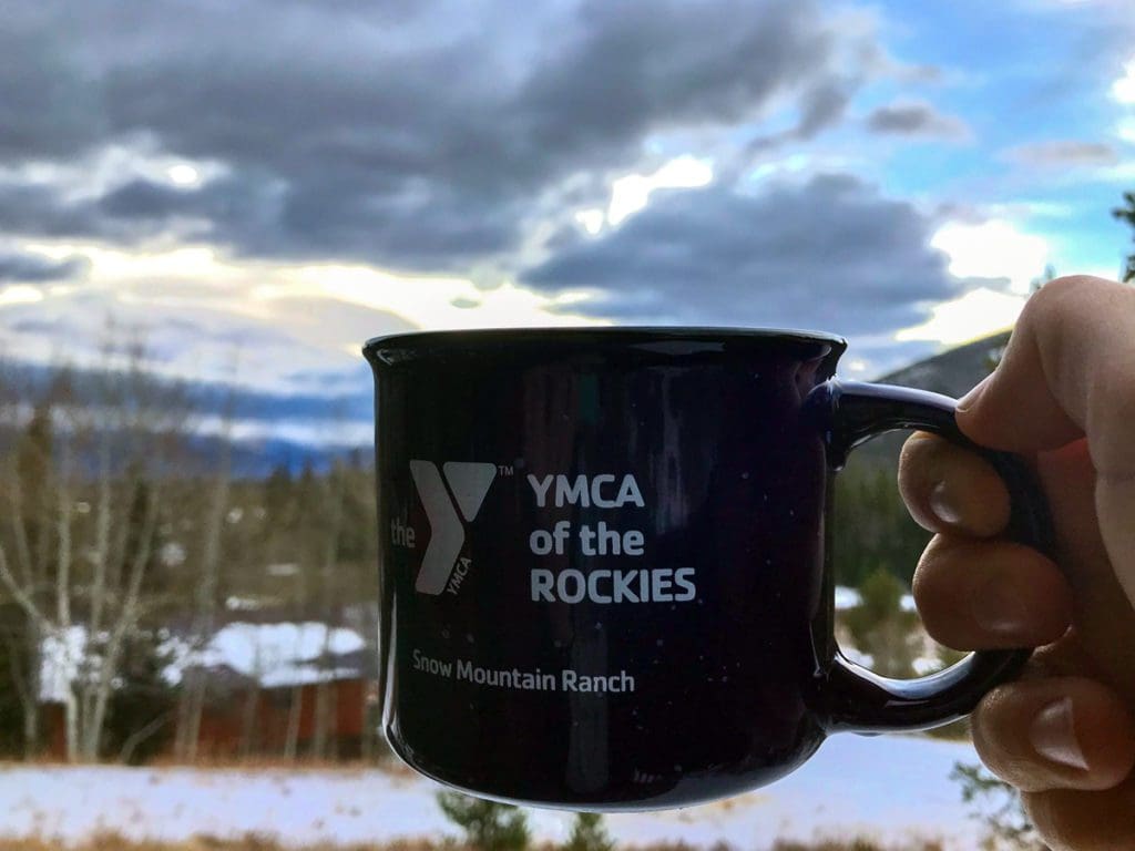 A hand holds a warm cup of hot cocoa, the mug reads "YMCA of the Rockies" with a scenic view in the distance.