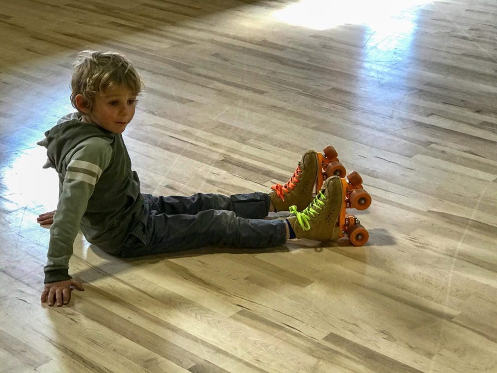 A young boy sits on a gym floor, wearing roller skates.