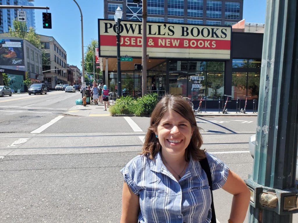 A woman stands on a street corder, with Powell's Books behind her.