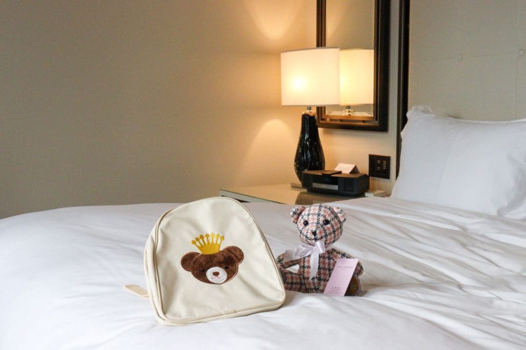 On top of a bed, rests a backpack and teddy bear from The Langham, Chicago.