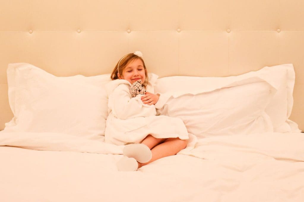 A young girl snuggles with a bear on a hotel bed, while staying at The Langham, Chicago.