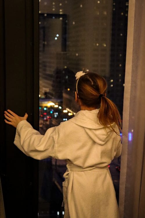 A young girl, wearing a bath robe, looks out a window onto a Chicago skyline at night.