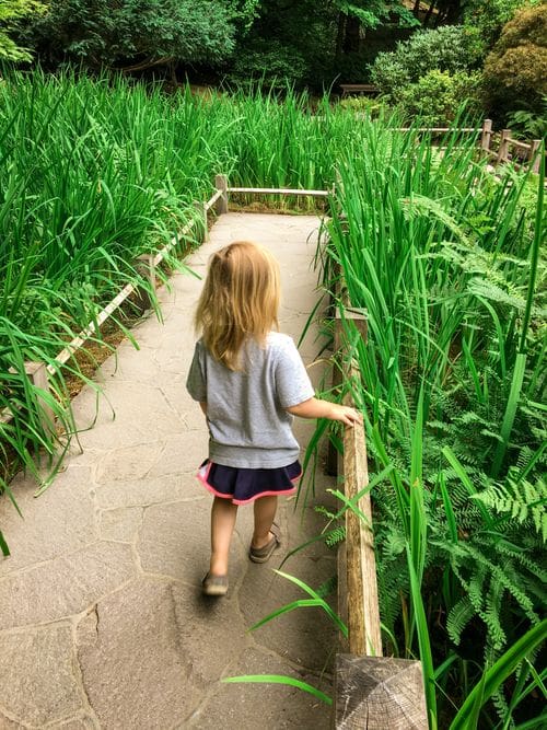 A young girl walks along a path, with green grasses on both sides, at the Japanese Garden, one of the best things to do in Portland with kids.