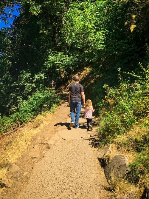 A dad holds his young daughter's hand as they hike along a dirt path.
