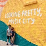 Two boys wearing sunglasses stand together in front of brightly colored street art in Nashville, reading "Looking Pretty, Music City".