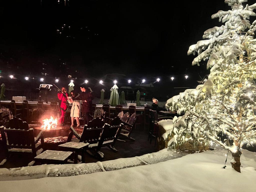 Several people sit and stand around a blazing outdoor fire pit, roasting marshmallows, at the Ritz-Carleton Bachelor Gulch.