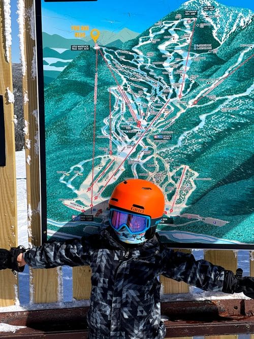 A young skier wearing an orange helmet stands in front of a slope map at Whiteface Mountain Ski Resort.