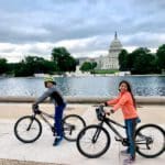 Two kids on bikes rest along the National Mall with monuments in the distance.